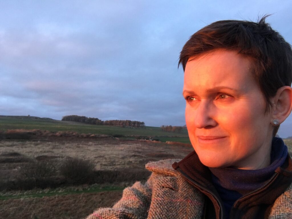 A close-up photo of Candida Meyrick outdoors in the countryside at sunset. The warm, golden light illuminates her face as she looks towards the horizon. She has short brown hair and is wearing a warm tweed coat against a backdrop of rolling hills under a dusky sky.